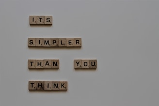 Wooden tiles with letters forming the phrase 'It's simpler than you think' are arranged on a neutral background. The tiles resemble those used in the game Scrabble, with each letter having a corresponding point value.