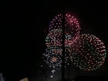 Bright and colorful fireworks illuminate the night sky with bursts of red, green, and pink lights. The explosions create intricate and symmetrical patterns that stand out against the dark background.