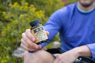 A person wearing a blue long-sleeved shirt is sitting outdoors, holding a bottle labeled as a hemp extract product. The background is filled with green foliage, suggesting a natural setting.