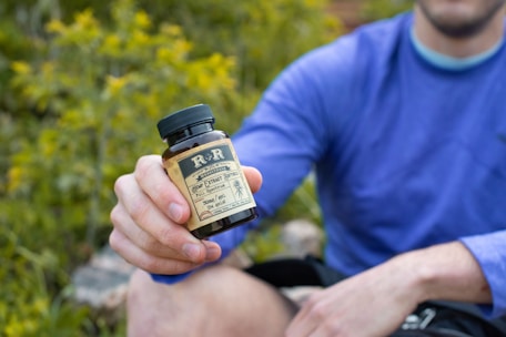A person wearing a blue long-sleeved shirt is sitting outdoors, holding a bottle labeled as a hemp extract product. The background is filled with green foliage, suggesting a natural setting.