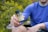 A person wearing a blue long-sleeved shirt is sitting outdoors, holding a bottle labeled as a hemp extract product. The background is filled with green foliage, suggesting a natural setting.
