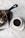 A cup of rich, dark coffee is placed on a white saucer next to a skillet with a dessert drizzled in chocolate sauce, accompanied by a few napkins on a wooden board. The background is a marbled table surface.