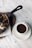 A cup of rich, dark coffee is placed on a white saucer next to a skillet with a dessert drizzled in chocolate sauce, accompanied by a few napkins on a wooden board. The background is a marbled table surface.