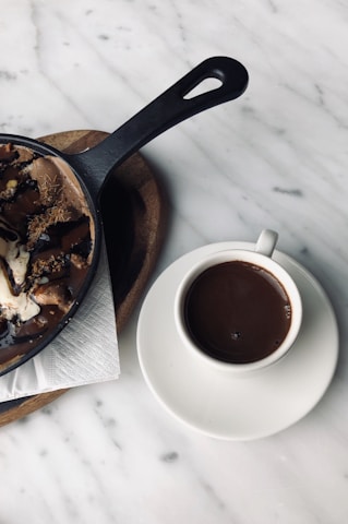 A cup of rich, dark coffee is placed on a white saucer next to a skillet with a dessert drizzled in chocolate sauce, accompanied by a few napkins on a wooden board. The background is a marbled table surface.