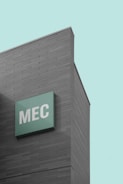 A modern building with a minimalist design features a large green sign displaying the letters 'MEC'. The facade is composed of horizontal gray panels, and the sky in the background is a clean, pastel blue.