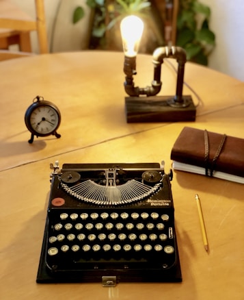 A vintage typewriter sits on a wooden table, accompanied by a small round clock, an Edison bulb lamp with a pipe-like structure, and a closed leather-bound notebook with a pencil beside it. The warm lighting and scattered objects suggest a cozy, nostalgic setting.