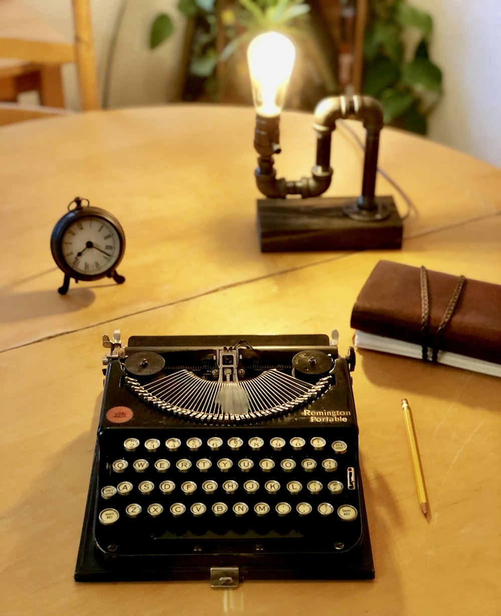 black typewriter machine beside yellow pencil, brown book, alarm clock and steam punk desk lamp on table