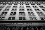 A black and white photograph of a tall, multi-story building with a symmetrical window arrangement. The facade features classical architectural elements and the words 'General Buildings' are prominently displayed at the base.