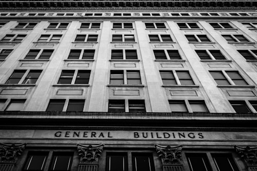 A black and white photograph of a tall, multi-story building with a symmetrical window arrangement. The facade features classical architectural elements and the words 'General Buildings' are prominently displayed at the base.