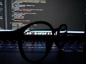 Spectacles are placed on a laptop keyboard, with a computer screen displaying code in the background.