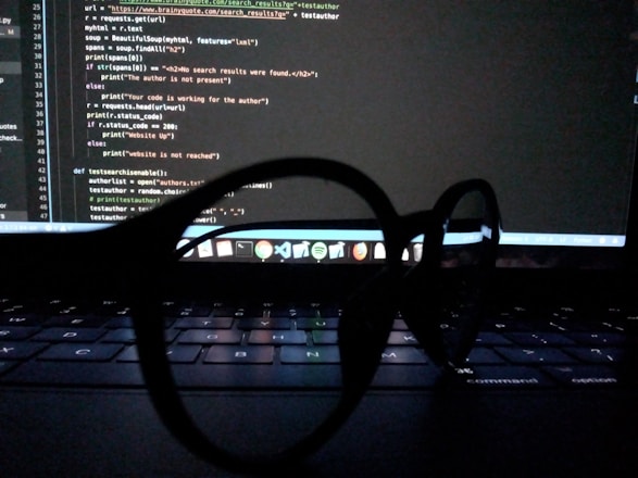 Spectacles are placed on a laptop keyboard, with a computer screen displaying code in the background.