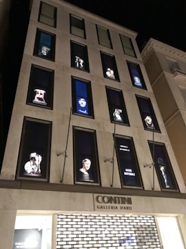 A multi-story building at night featuring large windows with illuminated art pieces. Each window displays a distinct artwork, including sculptures and portraits with a focus on modern and classical themes. The bottom of the building has a sign reading 'CONTINI Galleria D'Arte'.