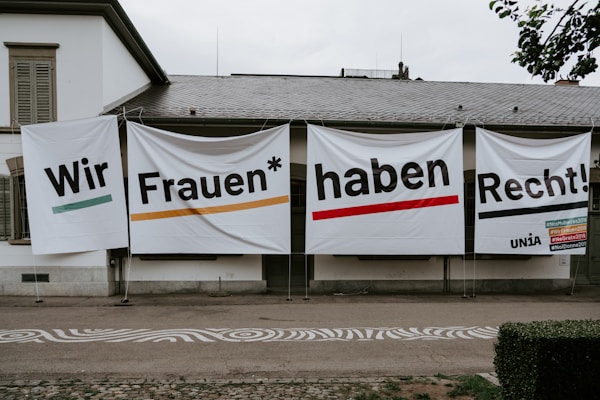 Large banners with bold text hang on the exterior wall of a building. The text is in German and translates to 'We women have rights!'. Several hashtags and the name 'UNIA' are visible on the right banner.