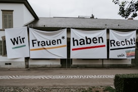 Large banners with bold text hang on the exterior wall of a building. The text is in German and translates to 'We women have rights!'. Several hashtags and the name 'UNIA' are visible on the right banner.