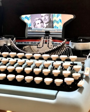 A vintage typewriter with creamy white keys sits prominently in the foreground. The typewriter's carriage is adorned with a retro, patterned background featuring a black and white photograph of a man and a woman. The image evokes a sense of nostalgia with its classic design and elements.