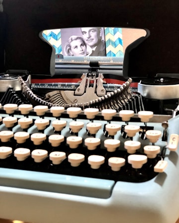 A vintage typewriter with creamy white keys sits prominently in the foreground. The typewriter's carriage is adorned with a retro, patterned background featuring a black and white photograph of a man and a woman. The image evokes a sense of nostalgia with its classic design and elements.