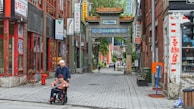 A narrow street scene in an urban Chinatown area features traditional Chinese architectural elements and signage. The image includes two people in the foreground, with one pushing the other in a wheelchair. The street is lined with brick buildings adorned with Asian-themed advertisements and banners. In the distance, a traditional Chinese gateway with ornate roofing is visible, adding cultural flair to the city environment.