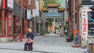A narrow street scene in an urban Chinatown area features traditional Chinese architectural elements and signage. The image includes two people in the foreground, with one pushing the other in a wheelchair. The street is lined with brick buildings adorned with Asian-themed advertisements and banners. In the distance, a traditional Chinese gateway with ornate roofing is visible, adding cultural flair to the city environment.