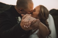 A close-up of a couple kissing, with the focus on their intertwined hands. The woman is wearing a wedding dress and a ring is visible on her finger. The background is softly blurred, suggesting a romantic outdoor setting.