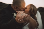 A close-up of a couple kissing, with the focus on their intertwined hands. The woman is wearing a wedding dress and a ring is visible on her finger. The background is softly blurred, suggesting a romantic outdoor setting.