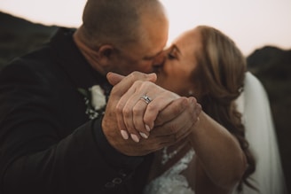 A close-up of a couple kissing, with the focus on their intertwined hands. The woman is wearing a wedding dress and a ring is visible on her finger. The background is softly blurred, suggesting a romantic outdoor setting.