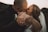 A close-up of a couple kissing, with the focus on their intertwined hands. The woman is wearing a wedding dress and a ring is visible on her finger. The background is softly blurred, suggesting a romantic outdoor setting.