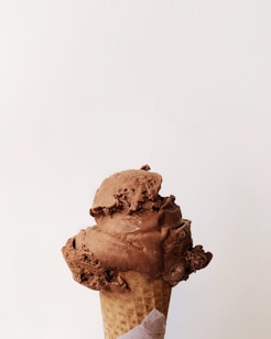 A close-up view of a chocolate ice cream cone with two scoops. The ice cream appears creamy and slightly melting, atop a waffle cone. The background is a plain, light color, which highlights the texture and richness of the chocolate ice cream.