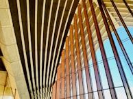 An architectural structure features a series of parallel concrete beams and large glass panels. The design emphasizes geometric patterns, with the beams casting shadows and the glass reflecting light. The setting suggests modernity and openness.