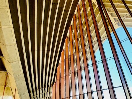 An architectural structure features a series of parallel concrete beams and large glass panels. The design emphasizes geometric patterns, with the beams casting shadows and the glass reflecting light. The setting suggests modernity and openness.