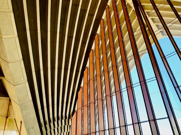 An architectural structure features a series of parallel concrete beams and large glass panels. The design emphasizes geometric patterns, with the beams casting shadows and the glass reflecting light. The setting suggests modernity and openness.