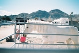 A white horse with braided mane stands in an outdoor pen, with a red bucket nearby. The setting is a ranch-like area with mountains in the background and a clear blue sky. The area is enclosed by metal fencing, and there are buildings and trailers visible.