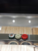 Three bottle caps are lined up on a shiny metallic surface. The caps are from different brands: a green Heineken cap with a red star, a pale green Birra Moretti cap with text indicating 'Limone 2% vol,' and a red Ichnusa non filtrata cap. The background is blurred and features light wooden tones.