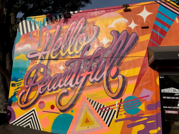 A vibrant and colorful mural with the large, stylized text 'Hello Beautiful' displayed across it. The background is filled with a variety of geometric shapes and patterns, including stripes, triangles, and circles. The colors are bright and bold, featuring elements like clouds and stars, creating a dynamic and cheerful composition.