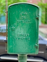 A green metal box with embossed text displaying 'City of Chicago Street Lighting' is mounted on a pole. The background is slightly blurred, showing foliage and part of a parked car.