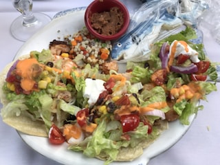 A plate of fish tacos topped with fresh lettuce, diced tomatoes, corn, black beans, and drizzled with an orange sauce. Next to the tacos, a red cup contains refried beans, and there is a side of couscous salad with vegetables. Plastic-wrapped cookies shaped like fish are also visible on the plate.