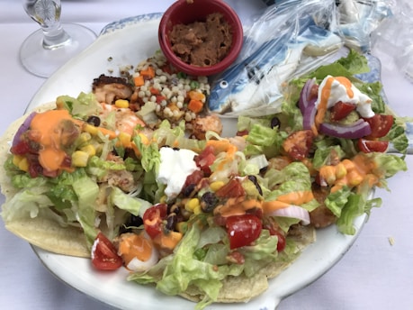 A plate of fish tacos topped with fresh lettuce, diced tomatoes, corn, black beans, and drizzled with an orange sauce. Next to the tacos, a red cup contains refried beans, and there is a side of couscous salad with vegetables. Plastic-wrapped cookies shaped like fish are also visible on the plate.