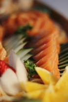 A close-up image of neatly arranged salmon sashimi slices, garnished with fresh parsley, lemon slices, and a radish flower. The presentation is accented by the soft focus background.