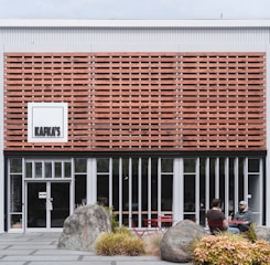 A modern building façade with a striking wood slat design features large vertical windows on the ground floor. Two people sit at a small outdoor table set surrounded by large rocks and decorative greenery, creating a relaxed atmosphere.