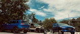 A serene outdoor scene featuring two parked cars, a blue SUV and a beige off-road vehicle, alongside another blue car with a visible hood scoop. The cars are positioned in a parking lot, surrounded by lush green trees under a partly cloudy blue sky.