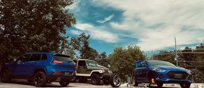 A serene outdoor scene featuring two parked cars, a blue SUV and a beige off-road vehicle, alongside another blue car with a visible hood scoop. The cars are positioned in a parking lot, surrounded by lush green trees under a partly cloudy blue sky.