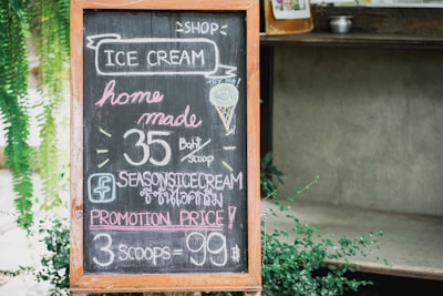 A wooden-framed blackboard displays colorful chalk writing advertising ice cream. It features the phrases 'home made' and 'promotion price' in pastel colors. Prices are written for scoops of ice cream, illustrated with a small chalk drawing of an ice cream cone. Leaves and greenery are visible around the edges.