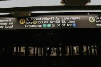 A subway station platform with signage displaying directions for various train lines, including the R, N, 4, 6, and E lines. The sign is well-lit against a dark background, indicating different routes and their respective platforms.
