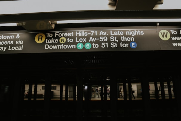 A subway station platform with signage displaying directions for various train lines, including the R, N, 4, 6, and E lines. The sign is well-lit against a dark background, indicating different routes and their respective platforms.