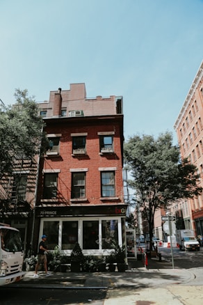 A red brick building located on a street corner with a small shop on the ground floor, surrounded by trees and a few pedestrians. The building has three stories with several windows, and there is signage for 'DIG' visible above the shop entrance. A truck is parked along the street, which is lined with potted plants.