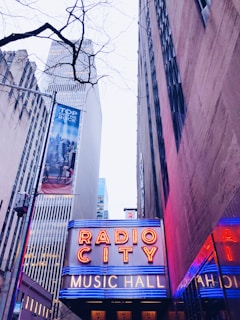 Neon lights illuminate the entrance of Radio City Music Hall. Surrounding skyscrapers stretch upwards, with a stark tree branch in the foreground. A cloudy sky looms overhead, and a billboard advertising 'Top of the Rock' is visible on a nearby building.