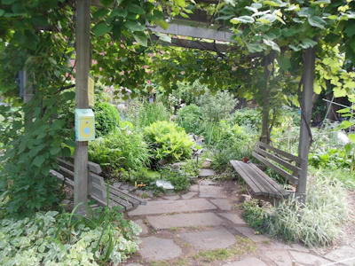 Community garden area with stone pathways and oak benches surrounded by greenery.