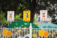 Three colorful signs with animated characters are displayed at the entrance of a school. The center sign is yellow with a blue boy holding a toy, while the two white signs on either side feature a girl in a red dress and yellow outfit. The signs are mounted on a fence decorated with floral motifs and playful designs, surrounded by greenery.