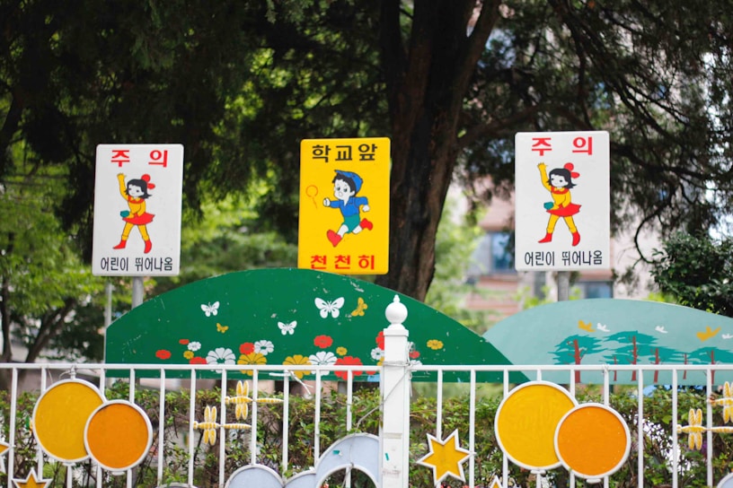 Three colorful signs with animated characters are displayed at the entrance of a school. The center sign is yellow with a blue boy holding a toy, while the two white signs on either side feature a girl in a red dress and yellow outfit. The signs are mounted on a fence decorated with floral motifs and playful designs, surrounded by greenery.