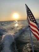 A coastal sunset with waves rolling in, silhouetted by a large American flag fluttering in the breeze.