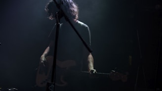 A musician stands in subdued lighting with a guitar, partially obscured by shadows and stage equipment. The scene conveys a moody, intimate atmosphere often associated with live music performances.